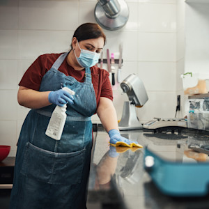A Hispanic woman uses cleaning chemicals to sanitize a food prep surface in a commercial kitchen.