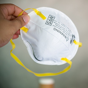 A person holds a white N95 respirator mask with yellow straps, commonly used for respiratory protection in dusty or hazardous environments.