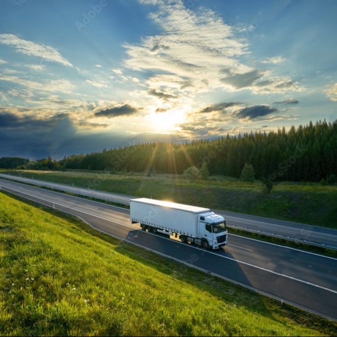 A white semi-truck travels along a multi-lane highway through a scenic countryside landscape at sunset.