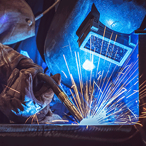 A professional welder in heavy protective gloves and helmet works with metal, causing bright sparks during the welding process.
