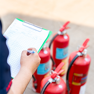 A safety inspector holds a clipboard and pen while recording details of several red fire extinguishers arranged on the ground during a safety inspection.