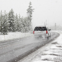 A white SUV travels along a winding road in heavy snowfall, with snow-covered trees lining the roadside and patches of snow on the ground.