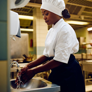 A Black female chef washes her hands at a sink in a commercial kitchen.