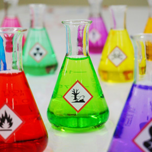 A collection of laboratory flasks filled with brightly colored liquids, each labeled with distinct chemical hazard symbols, arranged on a clean white surface in a scientific setting.