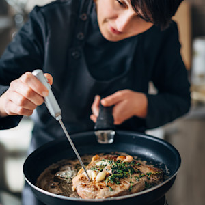 A caucasian female chef uses a meat thermometer on a steak cooking in a pan.