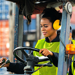 A woman operates a forklift in an industrial area, wearing a neon safety vest and yellow ear protection.