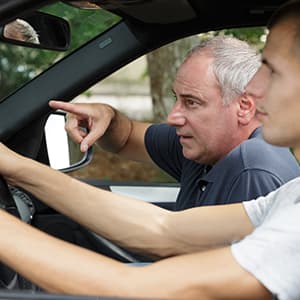 A driving instructor seated beside a student points toward the road while giving driving instructions inside a car.