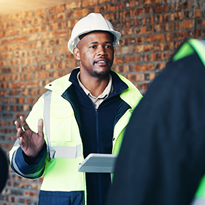 A construction professional wearing a hard hat and high-visibility vest holds a digital tablet while discussing work with a colleague at an indoor jobsite.