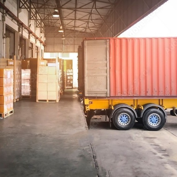 A shipping container trailer backed into a warehouse loading dock with pallets of goods prepared for loading or unloading.