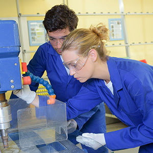 Two workers in blue lab coats and eye protection operate a drill press during hands-on safety or technical training.