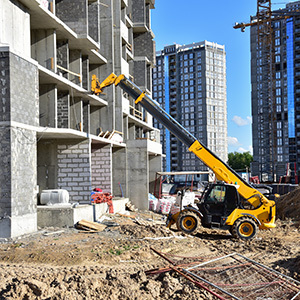 A yellow telescopic handler extends its boom to lift construction materials onto the upper level of a partially completed concrete building, with dirt ground, rebar and modern apartment buildings visible in the background.