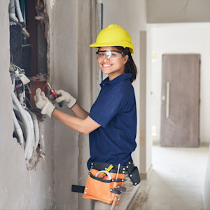 Portrait of smiling young female electrician using pliers while fixing wires in wall at incomplete house construction site.