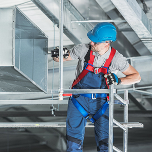 A construction worker in a hard hat, gloves and fall protection harness inspects industrial ductwork while standing on a scaffold at a jobsite.