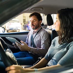 A driving instructor sits in the passenger seat and reviews information on a clipboard while discussing driving instruction with a student.