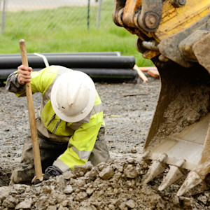 A construction worker wearing a hard hat and high-visibility jacket uses a shovel to dig a trench in rocky soil.