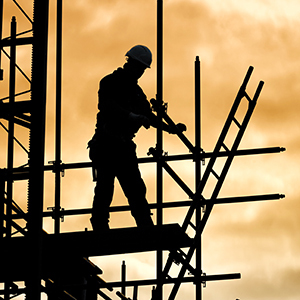 A silhouetted construction worker stands on a scaffold platform with ladders and guardrails visible against a warm sky.