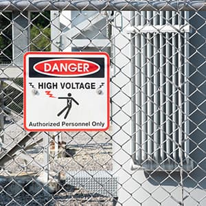 A warning sign reading “Danger High Voltage” is attached to a chain-link fence surrounding an electrical power facility.