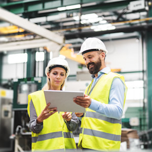 A male and female engineer wearing white safety helmets and yellow reflective vests stand in a modern industrial factory, reviewing information on a digital tablet while surrounded by large machinery.