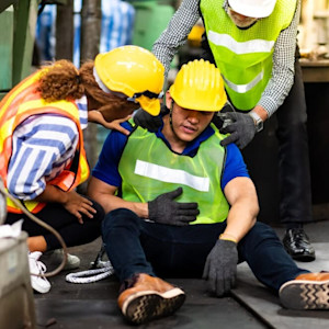 A male construction worker in a yellow hard hat and green safety vest sits on the ground clutching his chest, appearing injured. Two coworkers in safety gear kneel beside him, offering assistance.