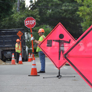 Two construction workers wearing orange helmets manage traffic around a road work area. Bright orange signs, including 'Work Area Ahead' and a flagger symbol, are displayed along with traffic cones.