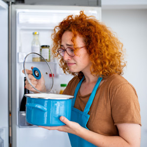 A red haired woman with a concerned expression checks a pot of food in the fridge.