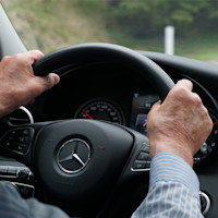 Close-up view of a person’s hands gripping the steering wheel of a Mercedes-Benz vehicle, with the dashboard and speedometer visible in the background.