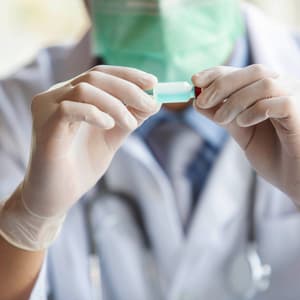 A laboratory professional wearing protective gloves examines a small sample tube in a laboratory setting.