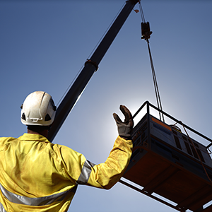 A construction signal person in protective gear directs a crane operator while a large suspended load is lifted against a clear sky.