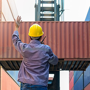 A worker wearing a hard hat signals while a crane or forklift moves a shipping container in an industrial yard.