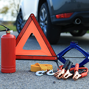 A roadside emergency kit is displayed in front of a car, including a red fire extinguisher, reflective warning triangle, yellow tow rope with hooks, jumper cables and a car jack placed on the pavement.