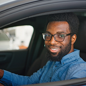 A man wearing glasses and a blue shirt sits in the driver’s seat of a car, smiling at the camera with his hand on the steering wheel.