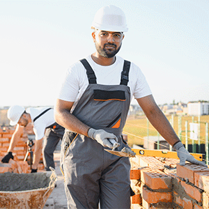 A construction worker wearing a white hard hat and gray overalls builds a brick wall using a trowel.