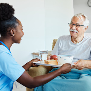 A female nursing home assistant presents a meal on a tray to an elderly man.