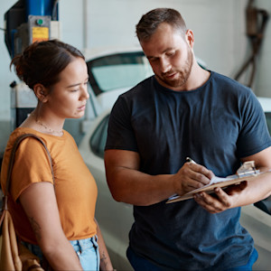 A man reviews an invoice on a clipboard with a woman at a car mechanic shop.