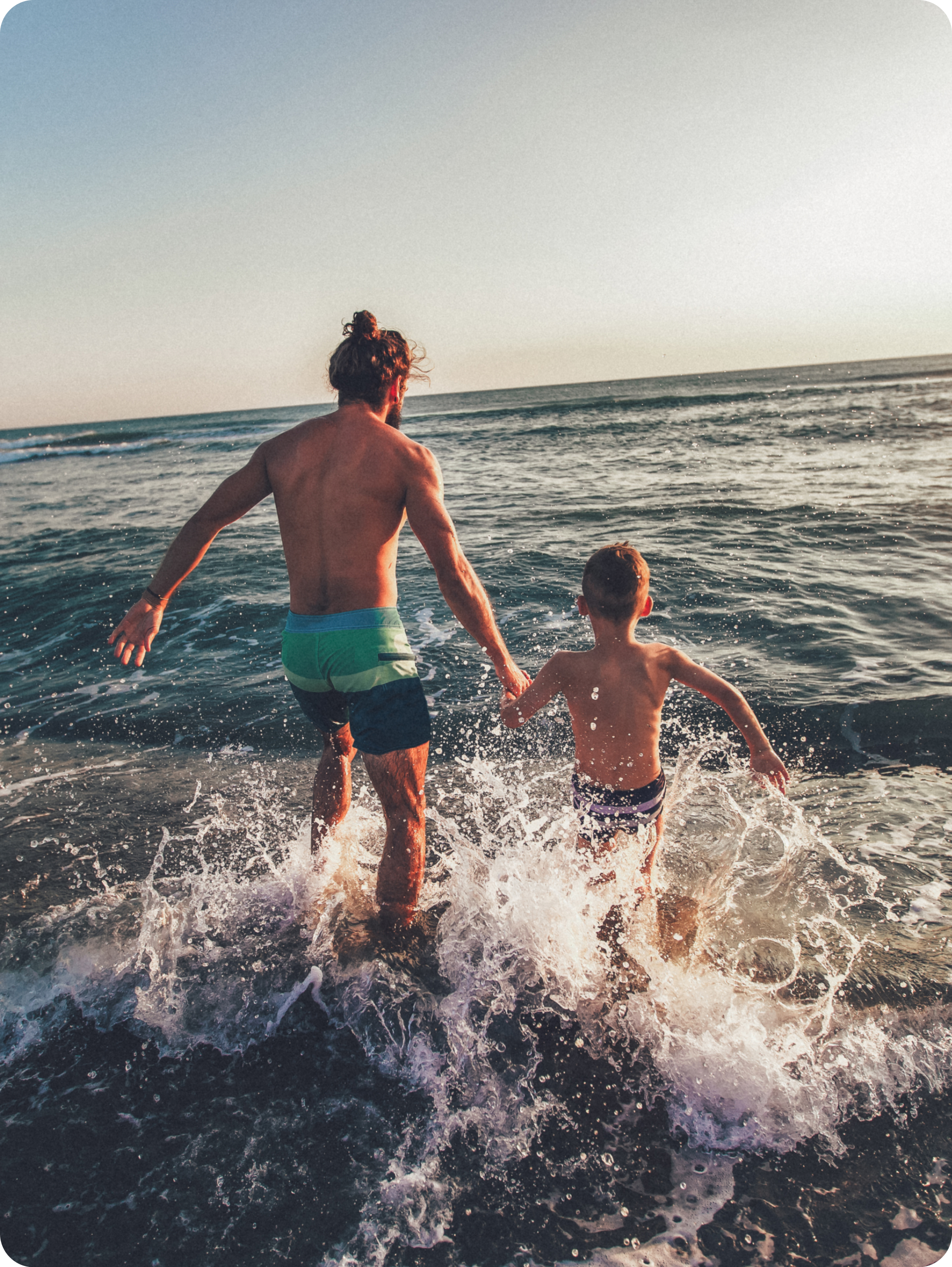 En far och dennes son springer över och igenom en liten våg i havet medans de håller händer