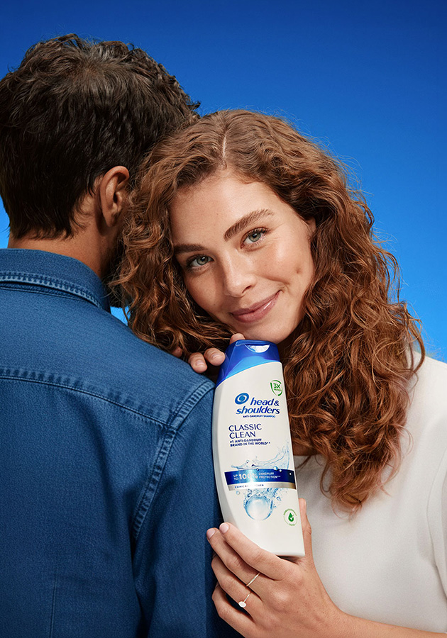 Eine junge Frau lehnt ihren Kopf an die Schulter eines Mannes und hält Head & Shoulders Classic Clean Shampoo in der Hand.
