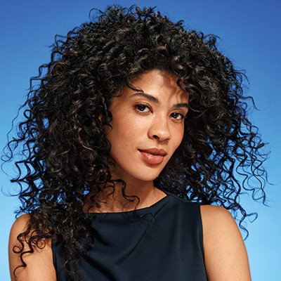 Lächelnde Frau mit langen, natürlich lockigen schwarzen Haaren vor blauem Hintergrund, die gesund aussehende und klar definierte Locken präsentiert.