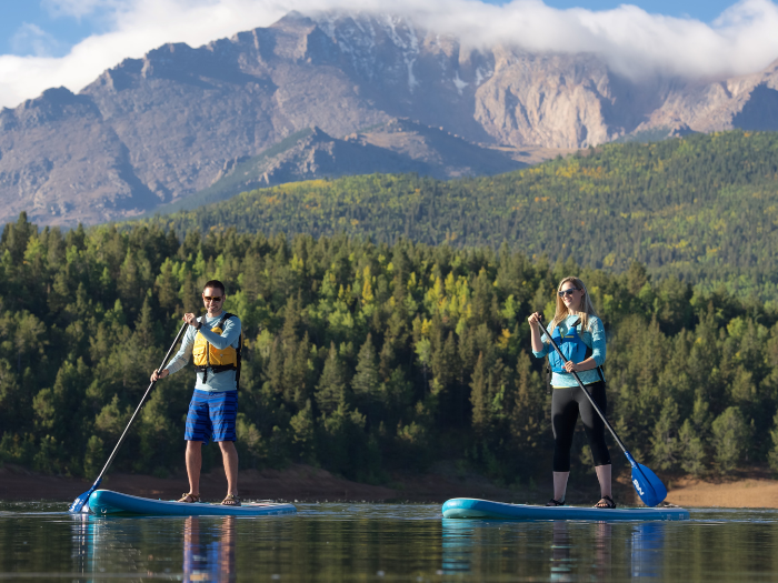 a younger male and female enjoying a leisurely paddleboard experience across a high mountain lake. Both are wearing lifejackets and smiling. The backdrop shows a forest of pine trees with tall mountains in the distance.