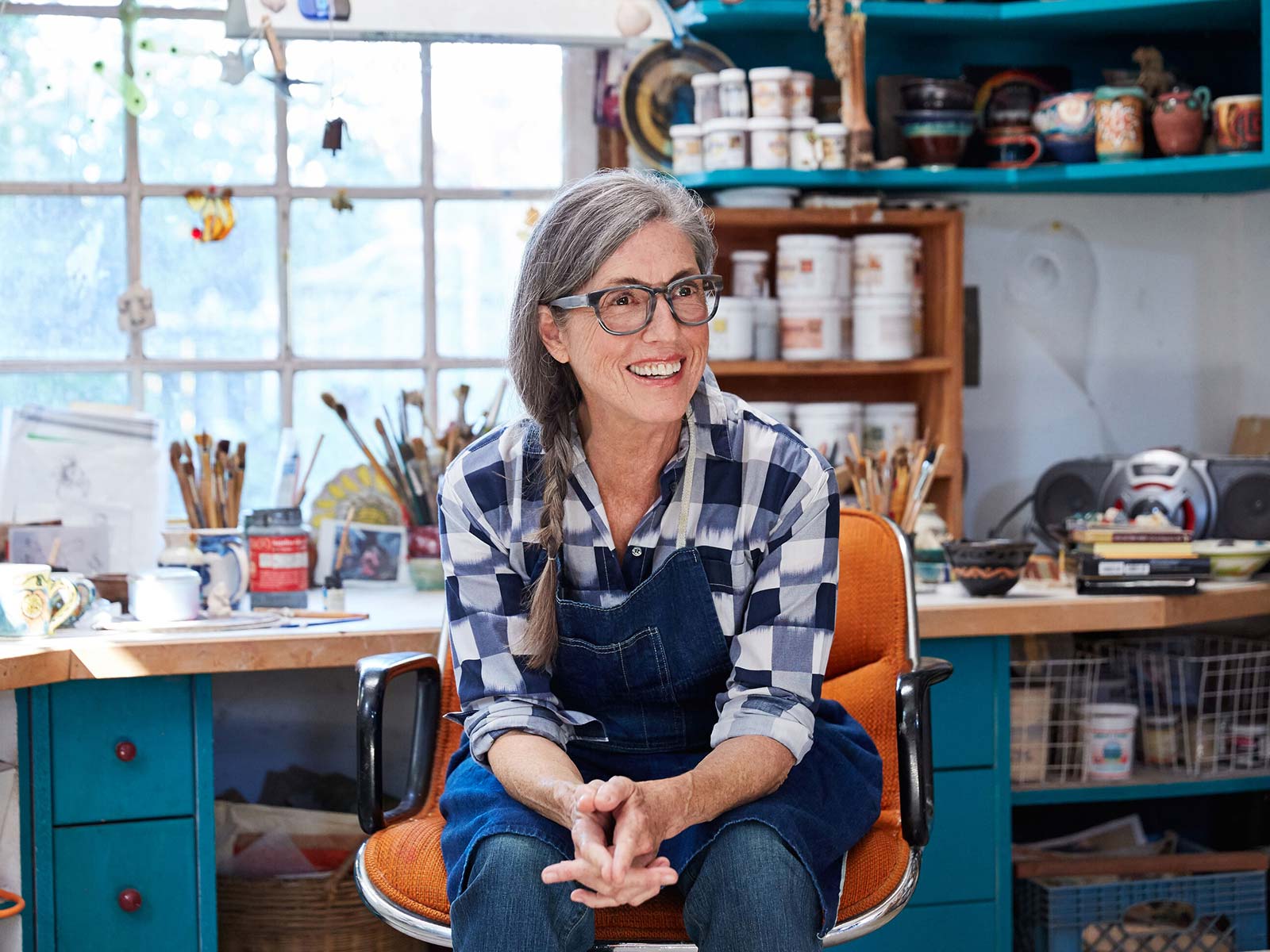 Female artist with gray side braid and glasses in a blue apron sitting in front of her studio workbench