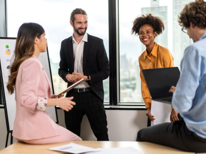 picture of four co-workers collaborating in a conference room with windows. The multi cultural image includes two women and two men. Two people (one female and one male) are sitting on a desk while the other two are standing with windows behind them that look out to a city scape.