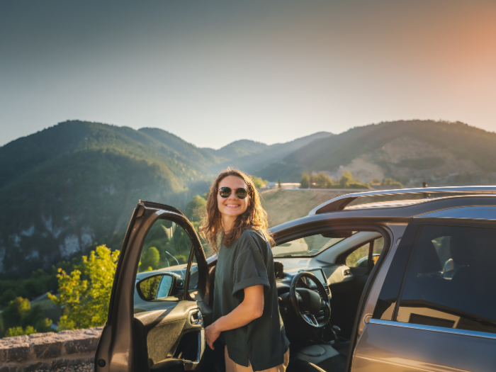 Mujer sonriente con gafas de sol saliendo de su coche estacionado en un mirador panorámico.