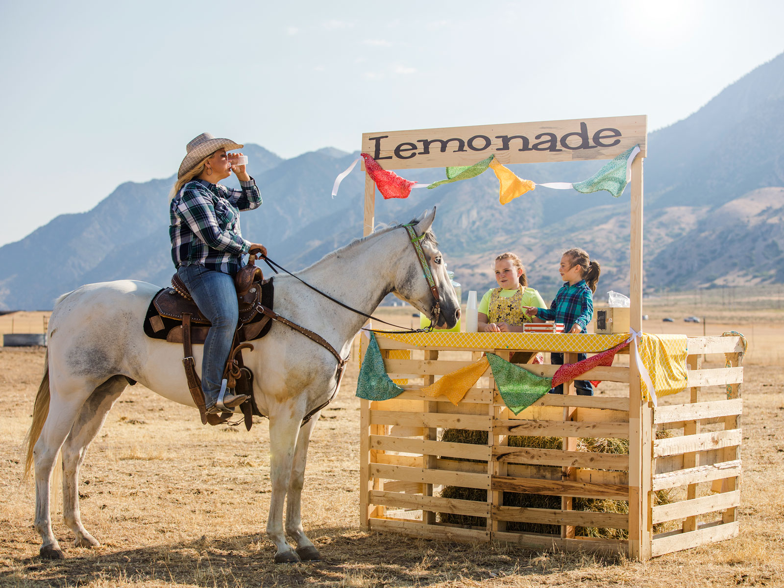 Una mujer en un caballo frente a un puesto de limonada con montañas de fondo