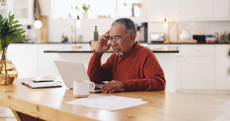 older minority male sitting at a kitchen table with a mug and a laptop looking worried.