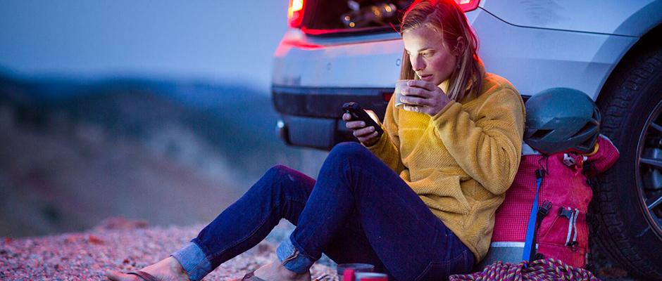Camping woman leans back on the bumper of her car, looking at her phone and sipping a hot beverage