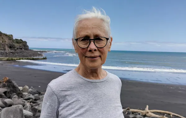 Old woman with white hair wearing glasses and a grey t-shirt, standing on a beach