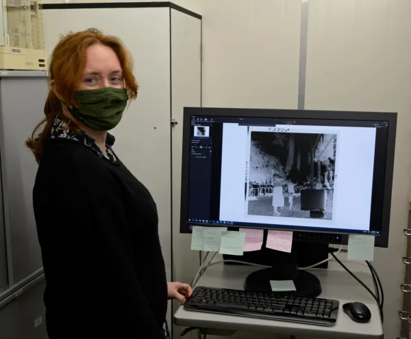 A woman with brown hair, wearing a black jacket is standing in front of a computer monitor displaying a black and white photo