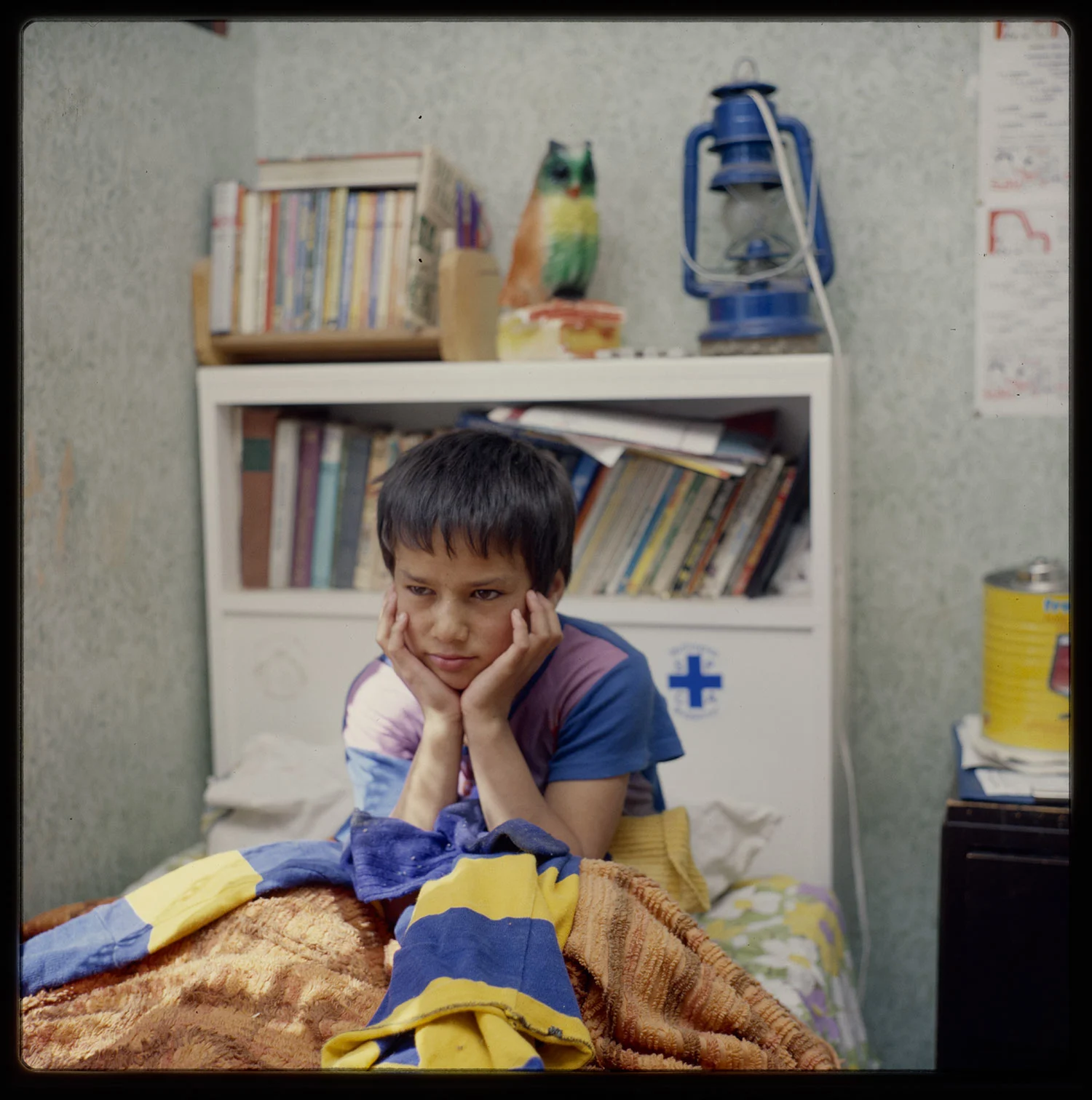A young boy sitting on a bed with his face resting on his two hands looking sad, a book shelf at the back