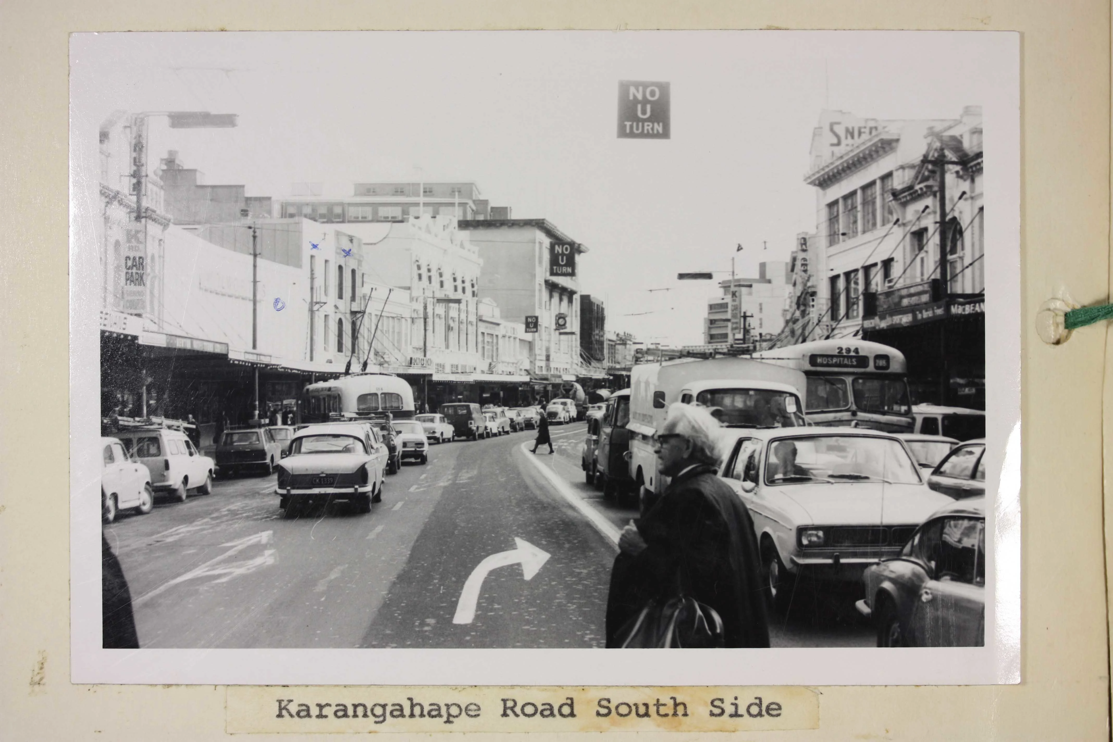Black and white image of a road with cars, buses and a man crossing it