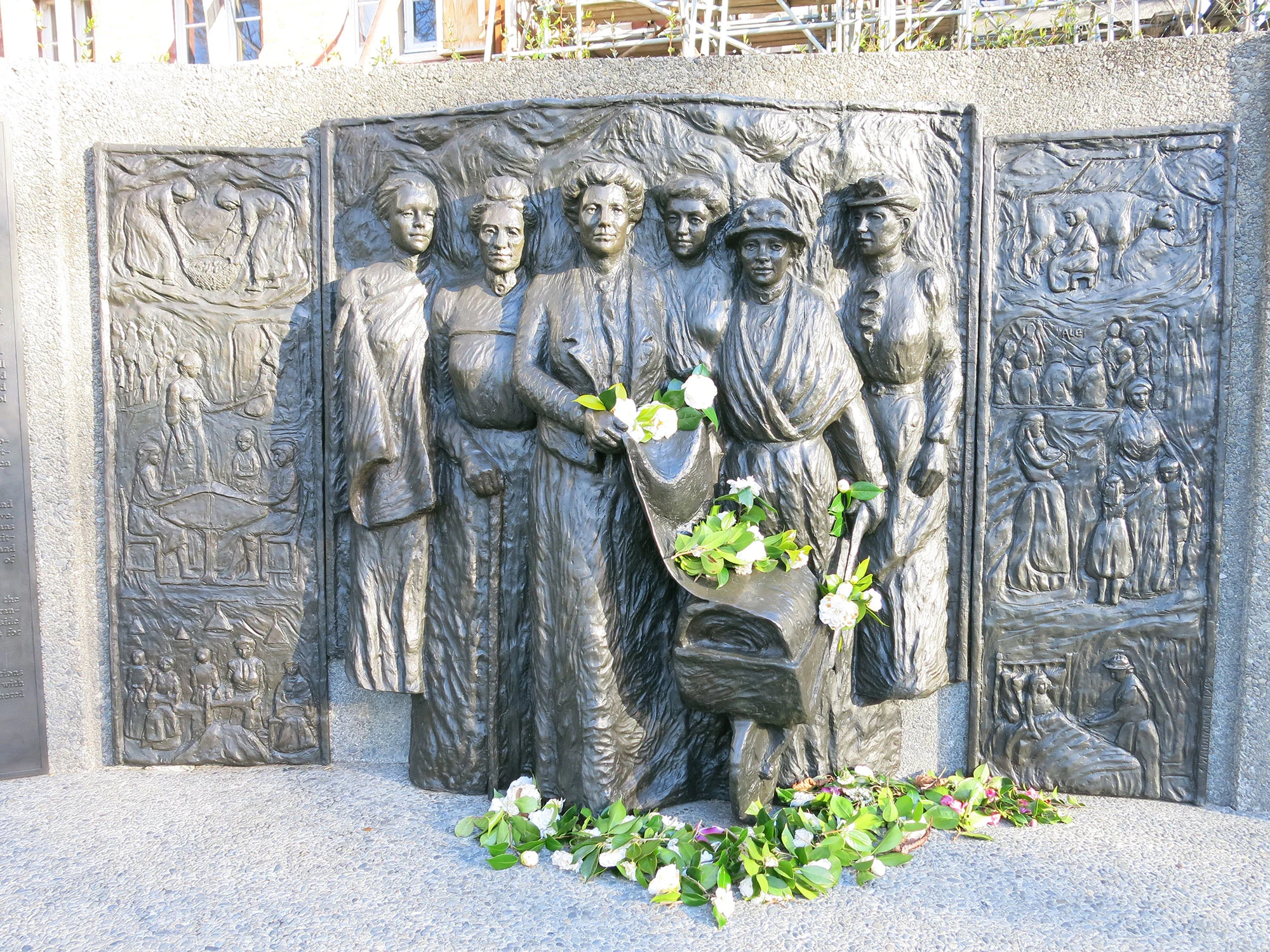 Statue on a wall of a group of women and green leaves in the front