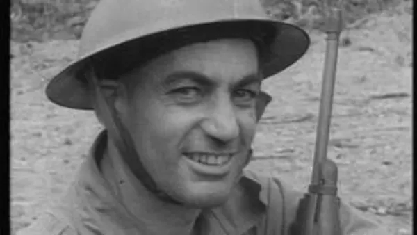 A black and white close up photo of a smiling man wearing a hard hat and holding a gun